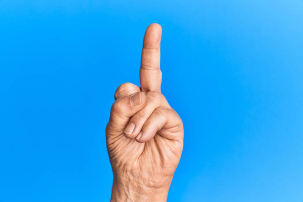 Hand eines älteren hispanischen Mannes über blauem isoliertem Hintergrund, der eine provokante und unhöfliche Geste zeigt, indem er mit dem Mittelfinger Fick-dich-Symbol macht  - Foto, Bild
