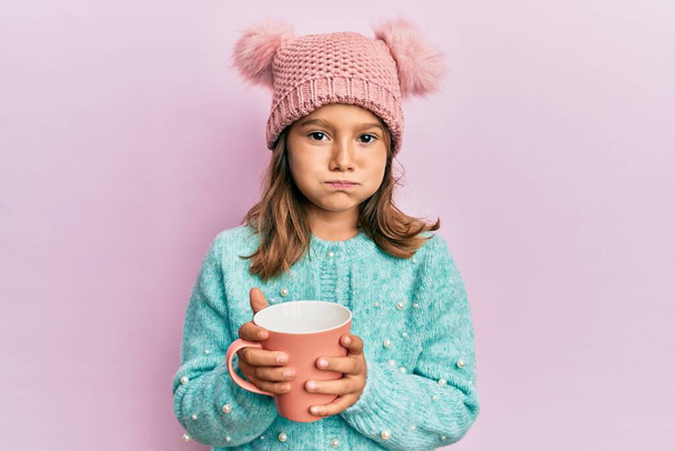 Маленькая красивая девочка с розовой кружкой в милой шерстяной шляпе пыхтя щеки с забавным лицом. рот надутый воздухом, ловящий воздух.  - Фото, изображение