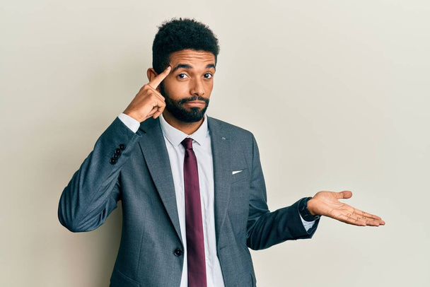 Handsome hispanic man with beard wearing business suit and tie confused and annoyed with open palm showing copy space and pointing finger to forehead. think about it.  - Photo, Image