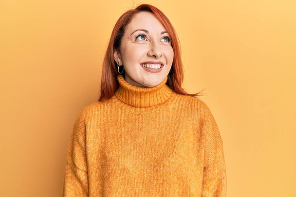 Mooie roodharige vrouw draagt casual winter trui over gele achtergrond kijken weg naar kant met glimlach op het gezicht, natuurlijke expressie. lachen vol vertrouwen.  - Foto, afbeelding