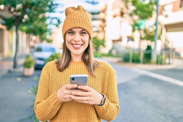 Nuori blondi nainen hymyilee onnellinen käyttäen älypuhelinta kaupungissa. - Valokuva, kuva