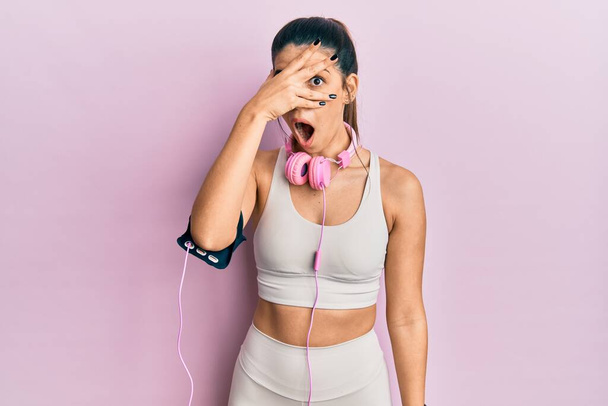 Young hispanic woman wearing gym clothes and using headphones peeking in shock covering face and eyes with hand, looking through fingers with embarrassed expression.  - Φωτογραφία, εικόνα