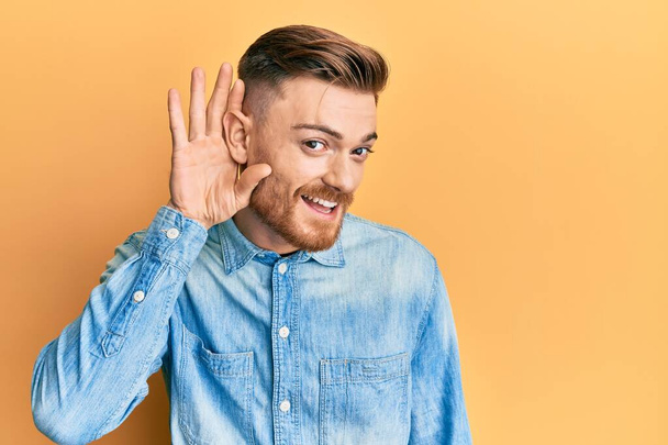Young redhead man wearing casual denim shirt smiling with hand over ear listening and hearing to rumor or gossip. deafness concept.  - Photo, Image