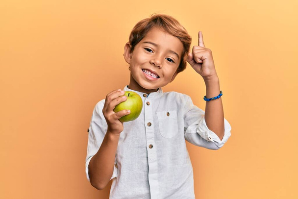 Чарівна латинська дитина тримає зелене яблуко посміхаючись з ідеєю або питанням, вказуючи пальцем щасливе обличчя, номер один
  - Фото, зображення