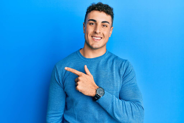 Hispanic young man wearing casual winter sweater cheerful with a smile on face pointing with hand and finger up to the side with happy and natural expression  - Photo, Image