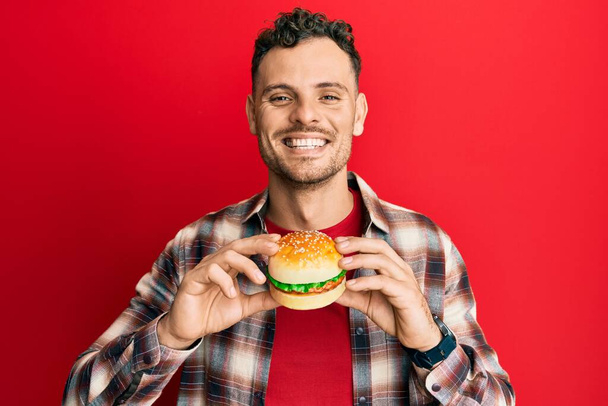 Lezzetli bir klasik burger yiyen genç İspanyol adam mutlu ve havalı bir gülümsemeyle gülümsüyor. diş gösterme.  - Fotoğraf, Görsel