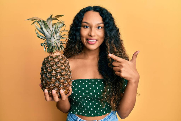 Fiatal latin nő kezében ananász mosolygós boldog mutató kéz és ujj  - Fotó, kép