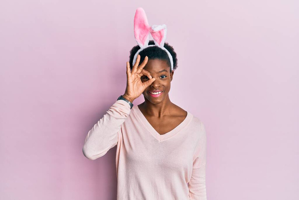 Young african american girl wearing cute easter bunny ears doing ok gesture with hand smiling, eye looking through fingers with happy face.  - Foto, afbeelding