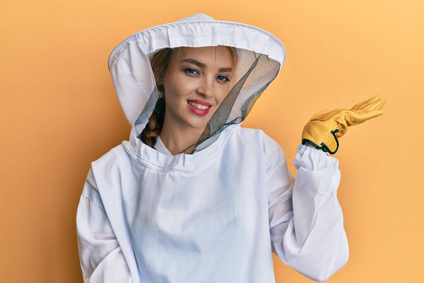 Beautiful blonde caucasian woman wearing protective beekeeper uniform smiling cheerful presenting and pointing with palm of hand looking at the camera.  - Valokuva, kuva