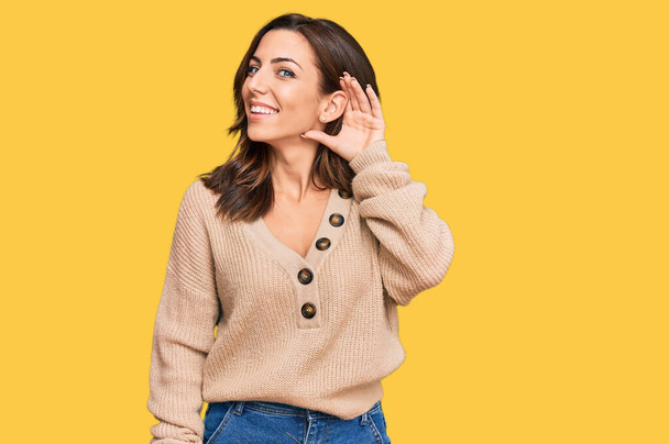 Jonge brunette vrouw draagt casual winter trui glimlachen met hand op oor luisteren naar een hoorzitting naar geruchten of roddels. doofheid.  - Foto, afbeelding