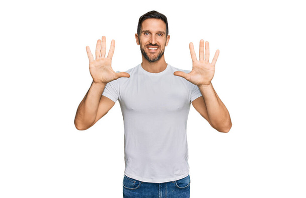 Hezký muž s plnovousem na sobě ležérní bílé tričko ukazující a ukazující nahoru s prsty číslo deset a usmívající se sebevědomě a šťastně.  - Fotografie, Obrázek