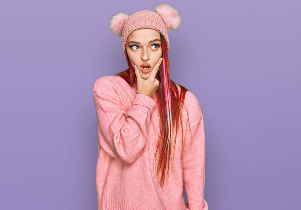 Young caucasian woman wearing casual clothes and wool cap looking fascinated with disbelief, surprise and amazed expression with hands on chin  - Foto, afbeelding