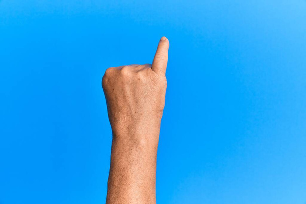 Ręka starszego Hiszpana nad niebieskim odizolowanym tłem pokazuje mały palec jako obietnicę, numer jeden.  - Zdjęcie, obraz