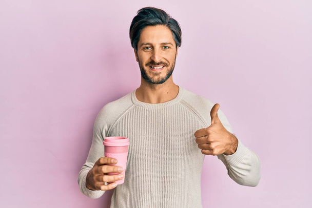 Young hispanic man drinking a take away cup of coffee smiling happy and positive, thumb up doing excellent and approval sign  - Photo, image
