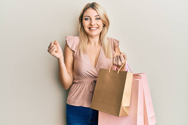 Genç beyaz kadın alışveriş torbaları tutuyor gururla bağırıyor, zaferi kutluyor ve başarıyı kollarını kaldırarak kutluyor.  - Fotoğraf, Görsel