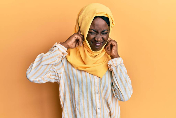 伝統的なイスラム教のヒジャーブスカーフを身に着けている美しいアフリカの若い女性は、大きな音楽のノイズのためのいらいらした表情で指で耳をカバーしています。聴覚障害の概念.  - 写真・画像