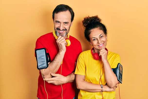 Μεσήλικες ζευγάρι Ισπανίδα γυναίκα και ο άνθρωπος φορώντας αθλητικά ρούχα και βραχιολάκι αναζητούν αυτοπεποίθηση στην κάμερα με χαμόγελο με σταυρωμένα χέρια και το χέρι έθεσε στο πηγούνι. θετική σκέψη.  - Φωτογραφία, εικόνα