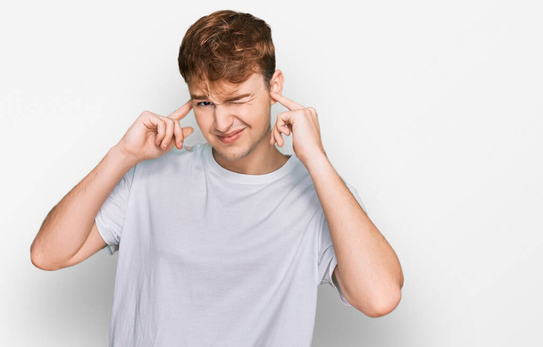 Jonge blanke man draagt casual wit t-shirt over oren met vingers met geïrriteerde uitdrukking voor het lawaai van luide muziek. doof concept.  - Foto, afbeelding