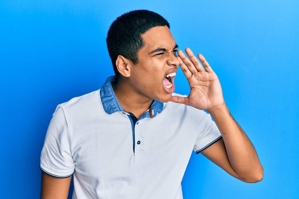 Jonge knappe Spaanse man in casual kleding schreeuwend en schreeuwend naast elkaar met hand op de mond. communicatieconcept.  - Foto, afbeelding