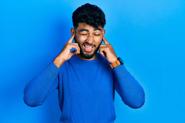 Arab man with beard wearing casual blue sweater covering ears with fingers with annoyed expression for the noise of loud music. deaf concept.  - Fotografie, Obrázek