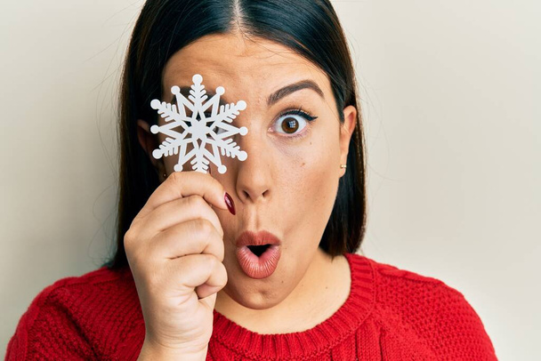 Beautiful brunette woman holding snowflake over eye scared and amazed with open mouth for surprise, disbelief face  - Photo, Image