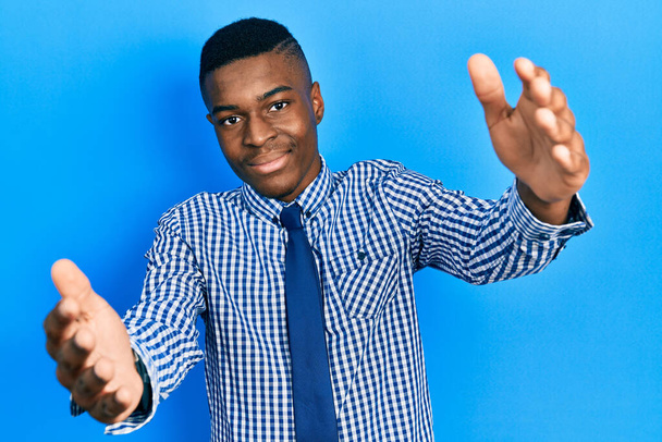 Młody Afroamerykanin w biznesowej koszuli i krawacie, patrzący w kamerę i uśmiechający się z otwartymi ramionami. radosna ekspresja obejmująca szczęście.  - Zdjęcie, obraz