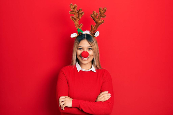Beautiful hispanic woman wearing deer christmas hat and red nose happy face smiling with crossed arms looking at the camera. positive person.  - Photo, Image