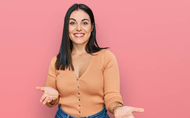 Молода іспаномовна жінка в повсякденному одязі усміхається весело з відкритими руками як дружні привітання, позитивні і впевнені привітання
  - Фото, зображення
