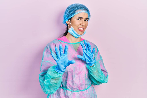 Young brunette woman wearing surgeon uniform and medical mask disgusted expression, displeased and fearful doing disgust face because aversion reaction. with hands raised  - Photo, Image