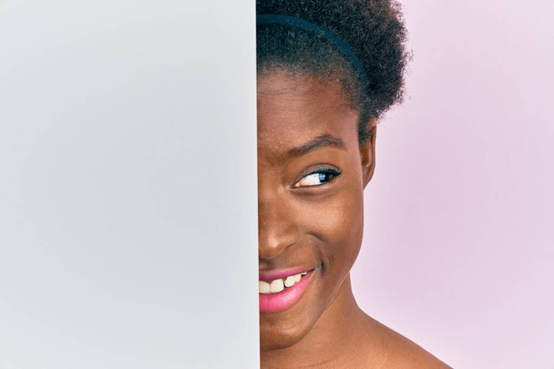 Młoda afrykańska dziewczyna trzyma pusty sztandar na pół twarzy uśmiechając się patrząc w bok i patrząc myśląc.  - Zdjęcie, obraz