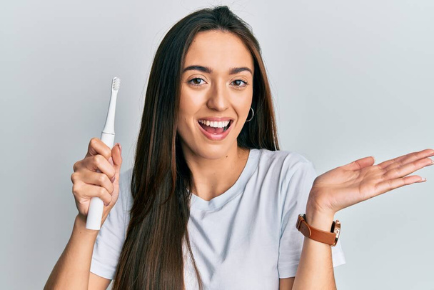 Joven chica hispana sosteniendo cepillo de dientes eléctrico celebrando logro con sonrisa feliz y expresión ganadora con la mano levantada  - Foto, Imagen