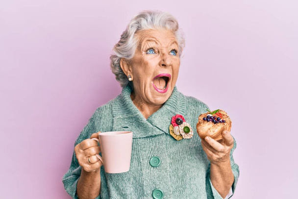 Ältere grauhaarige Frau trinkt eine Tasse Kaffee und isst Brötchen wütend und verrückt schreiend frustriert und wütend schreiend, schreiend vor Wut aufblickend.  - Foto, Bild