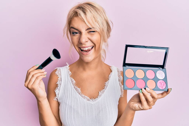 Young blonde girl holding makeup brush and blush winking looking at the camera with sexy expression, cheerful and happy face.  - Photo, Image