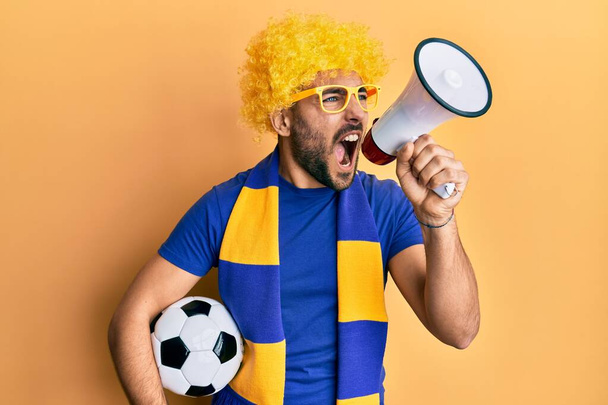 Young hispanic man football supporter holding soccer ball using megaphone angry and mad screaming frustrated and furious, shouting with anger. rage and aggressive concept.  - Zdjęcie, obraz