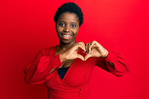 赤い背景の上にカジュアルな服を着てアフロの髪を持つ若いアフリカの女性は手で心のシンボル形状を行うの愛に笑みを浮かべて。ロマンチックなコンセプト.  - 写真・画像