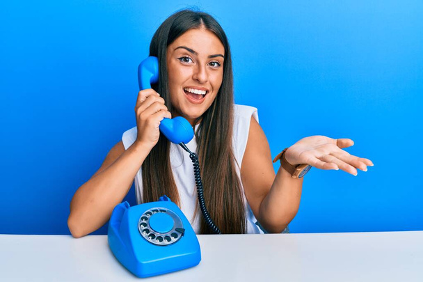 Beautiful hispanic woman speaking on vintage telephone celebrating achievement with happy smile and winner expression with raised hand  - Φωτογραφία, εικόνα