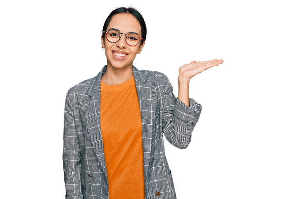 Junges hispanisches Mädchen in Businessjacke und Brille lächelt fröhlich und zeigt mit der Handfläche in die Kamera.  - Foto, Bild