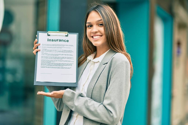 Młoda latynoska bizneswoman uśmiechnięta happy holding clipboard z umową ubezpieczenia w mieście. - Zdjęcie, obraz