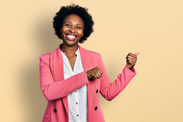 Αφροαμερικανή γυναίκα με αφρο μαλλιά που φοράει επαγγελματικό μπουφάν που δείχνει προς τα πίσω με το χέρι και τους αντίχειρες προς τα πάνω, χαμογελαστή αυτοπεποίθηση  - Φωτογραφία, εικόνα