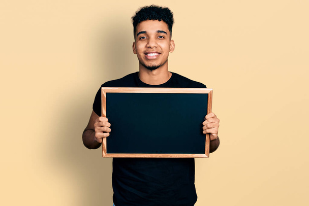 Young african american man holding blackboard looking positive and happy standing and smiling with a confident smile showing teeth  - Foto, Imagen