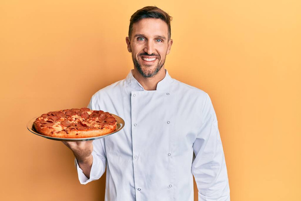 Hombre guapo con barba cocinero profesional sosteniendo pizza italiana mirando positiva y feliz de pie y sonriendo con una sonrisa confiada mostrando los dientes  - Foto, imagen