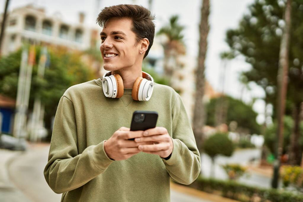 Nuori latino mies hymyilee onnellinen käyttäen älypuhelin ja kuulokkeet kaupungin. - Valokuva, kuva