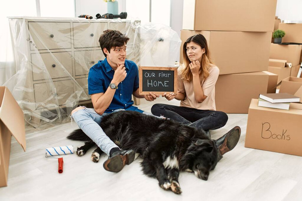 Jeune couple caucasien avec chien tenant notre premier tableau noir à la maison à la nouvelle maison pensant inquiet d'une question, préoccupé et nerveux avec la main sur le menton  - Photo, image