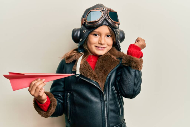 Kis gyönyörű lány visel pilóta egyenruhát tartó papír sík sikoltozik büszke, ünnepli győzelem és a siker nagyon izgatott felemelt karral  - Fotó, kép