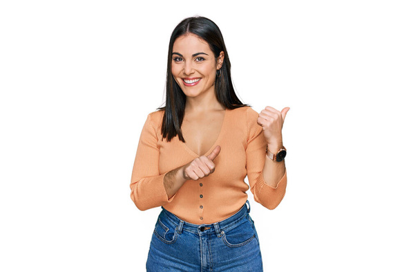 Young hispanic woman wearing casual clothes pointing to the back behind with hand and thumbs up, smiling confident  - 写真・画像