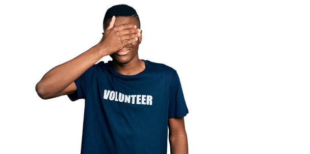 ボランティアのTシャツを着た若いアフリカ系アメリカ人の男性が笑顔で、驚きのために目を覆う顔を手で笑っています。視覚障害の概念.  - 写真・画像