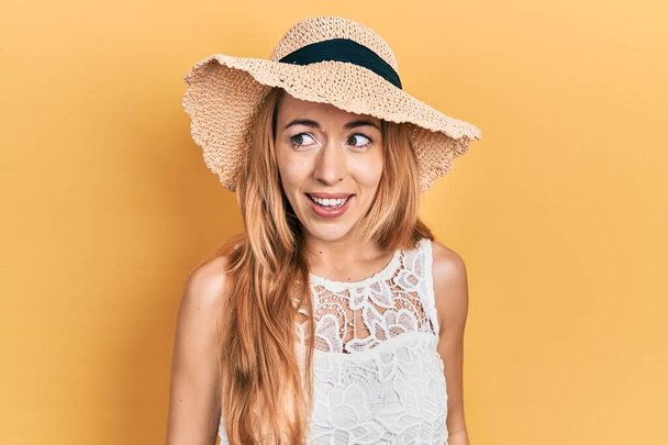 Jonge blanke vrouw met een zomerhoed die van de ene kant naar de andere kijkt met een glimlach op haar gezicht, natuurlijke uitdrukking. lachen vol vertrouwen.  - Foto, afbeelding