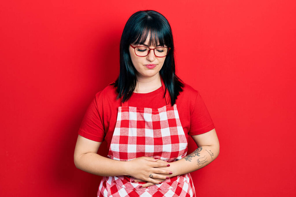Young hispanic woman wearing cook apron and glasses with hand on stomach because indigestion, painful illness feeling unwell. ache concept.  - Photo, Image