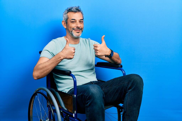 灰色の髪をしたハンサムな中年の男性が車椅子の成功サインに座って手で積極的なジェスチャーを行うと、親指を笑顔と幸せ。陽気な表情と勝者のジェスチャー.  - 写真・画像
