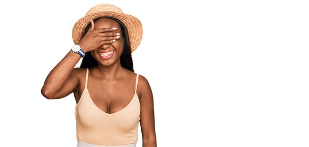 Egy fiatal fekete nő, aki nyári kalapot visel, mosolyog és nevetgél, miközben arcon takarja a szemeit meglepetésként. vak fogalom.  - Fotó, kép